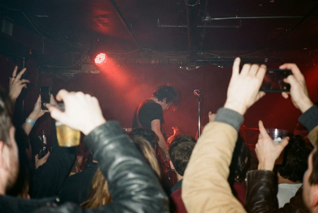 A guitarist's silhouette on a low stage seen over the shoulders of a packed standing crowd, red stage light and haze
